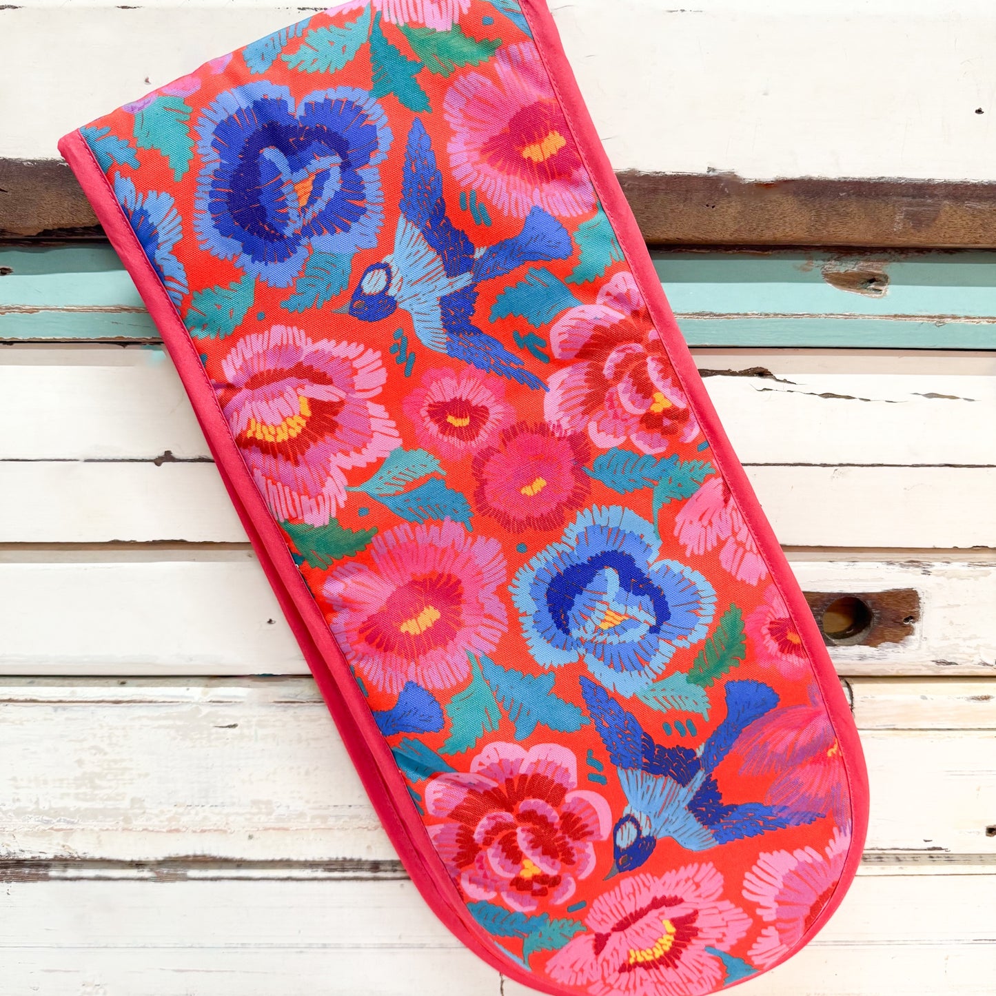 A rectangular quilted fabric item, likely a pillowcase or decorative cover, featuring a vibrant floral and bird pattern on a red-orange background. The design includes pink and blue flowers with green leaves and blue birds.