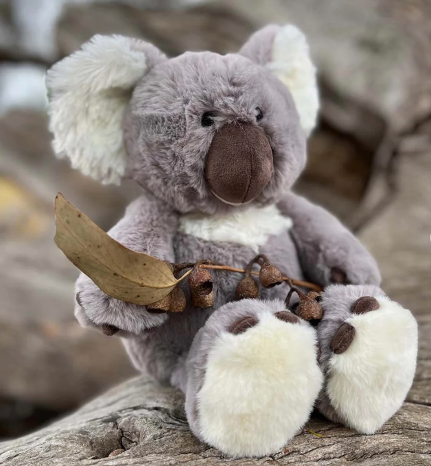 FURFOLK Koala Soft Toy. A plush toy koala softly sitting on a wooden surface, holding a green leaf, with a blurred background.