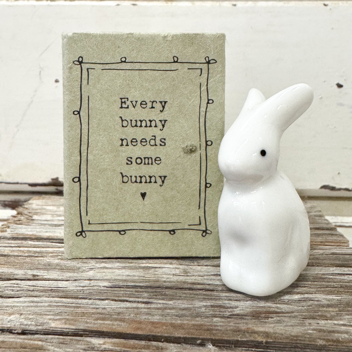 White ceramic bunny figurine next to a sign with text on a wooden surface Reads " Every Bunny needs some bunny"