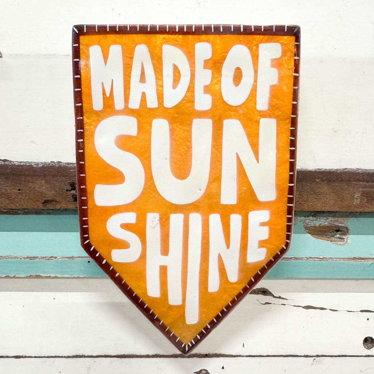 A shield-shaped decorative sign with the words “MADE OF SUN SHINE” in large white letters on a textured orange background, with a dark brown border, photographed against a distressed white and teal wooden surface.
