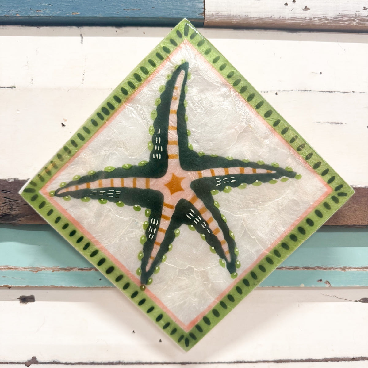 A square capiz shell tile with a diamond orientation, featuring a hand-painted starfish design in green, orange, and white, surrounded by a green dotted border on a rustic white wooden background.