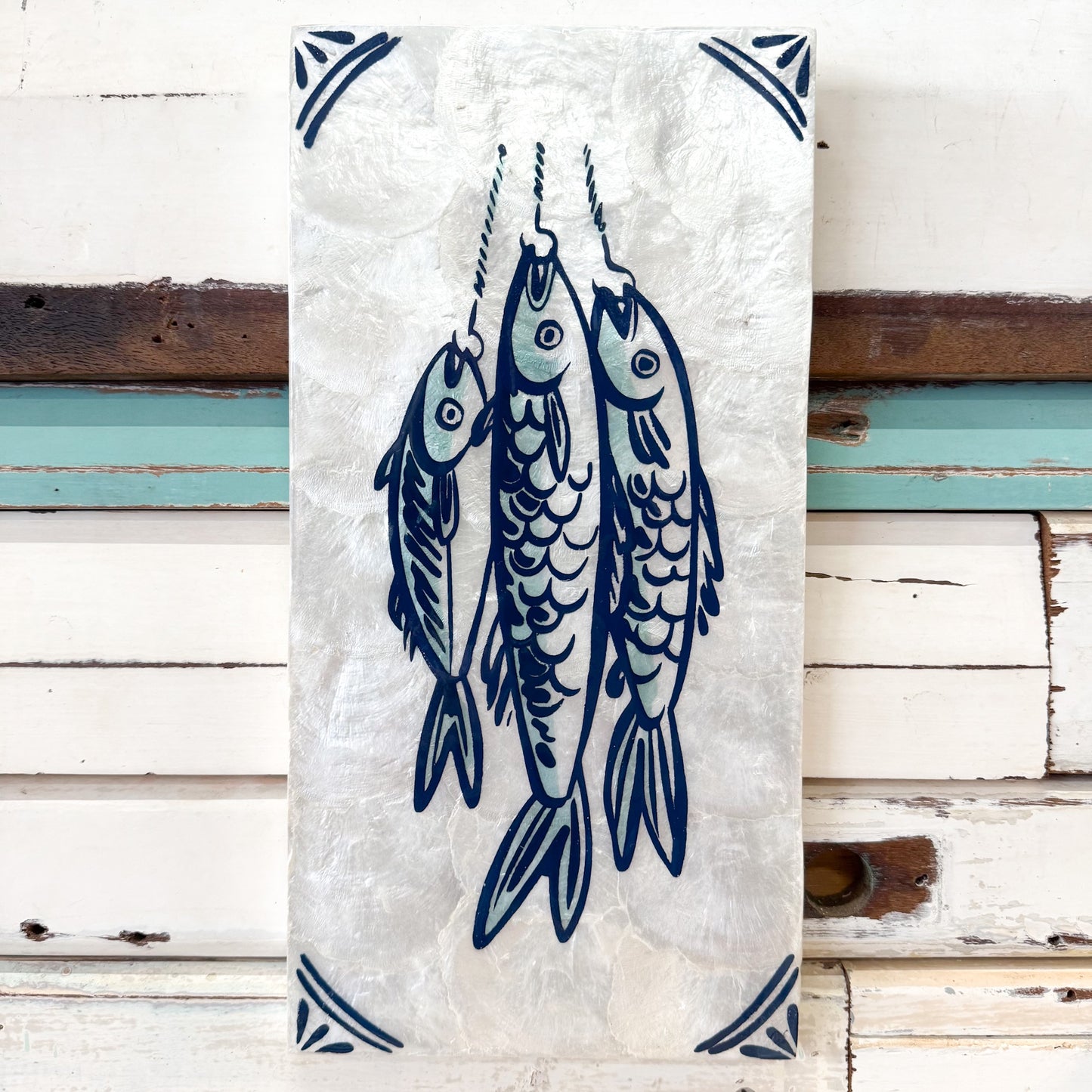 A rectangular white wall hanging or tile with a blue illustration of three fish hanging from hooks, bordered with blue corner motifs, displayed against a white wooden plank wall.
Jones & Co | Taverna Fish