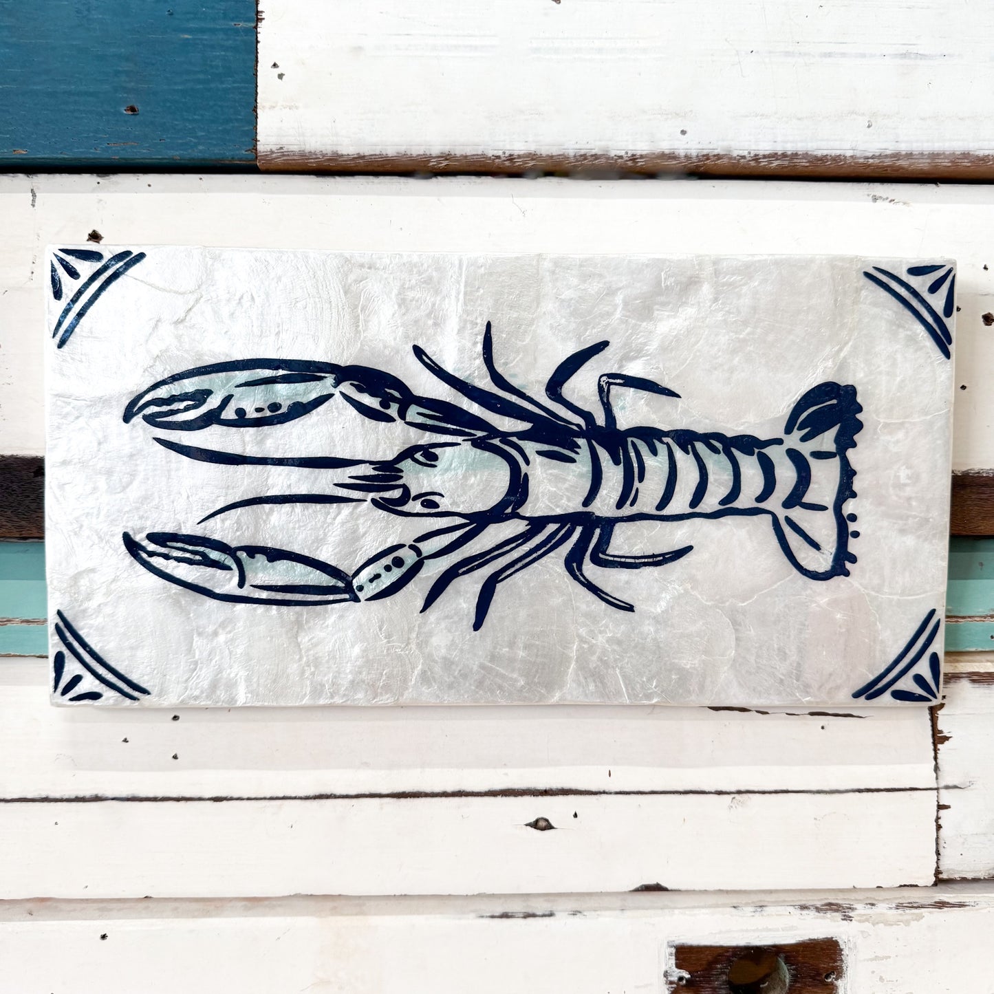 A rectangular decorative sign featuring a navy blue lobster illustration on a textured off-white background, mounted against a white wooden surface with teal and white painted boards.
Jones & Co | Taverna Lobster