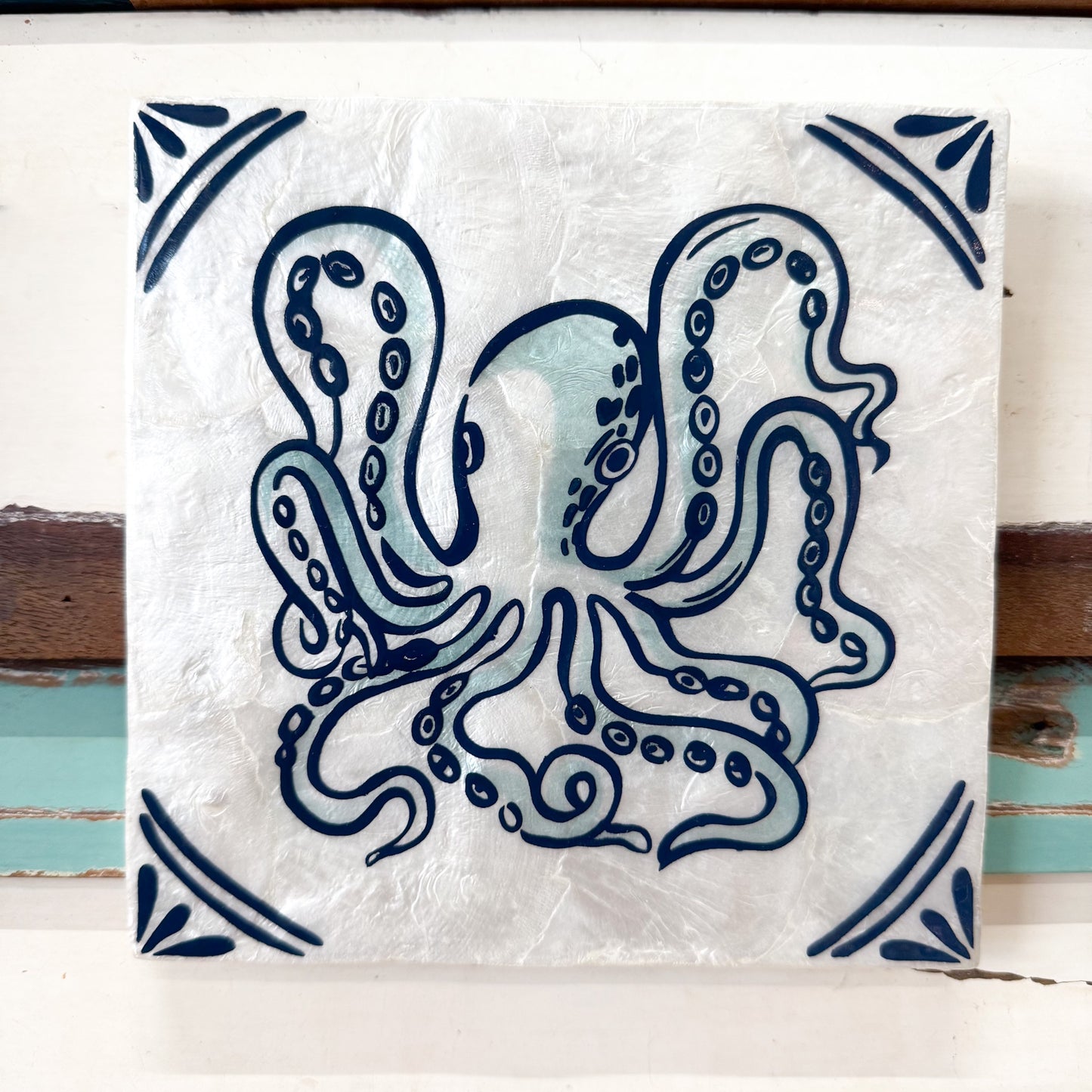 A square ceramic tile featuring a hand-painted octopus design in dark blue and light blue on a textured white background, with decorative corner motifs. The tile is displayed against a rustic white and brown wooden background. Jones & Co | Taverna Occy