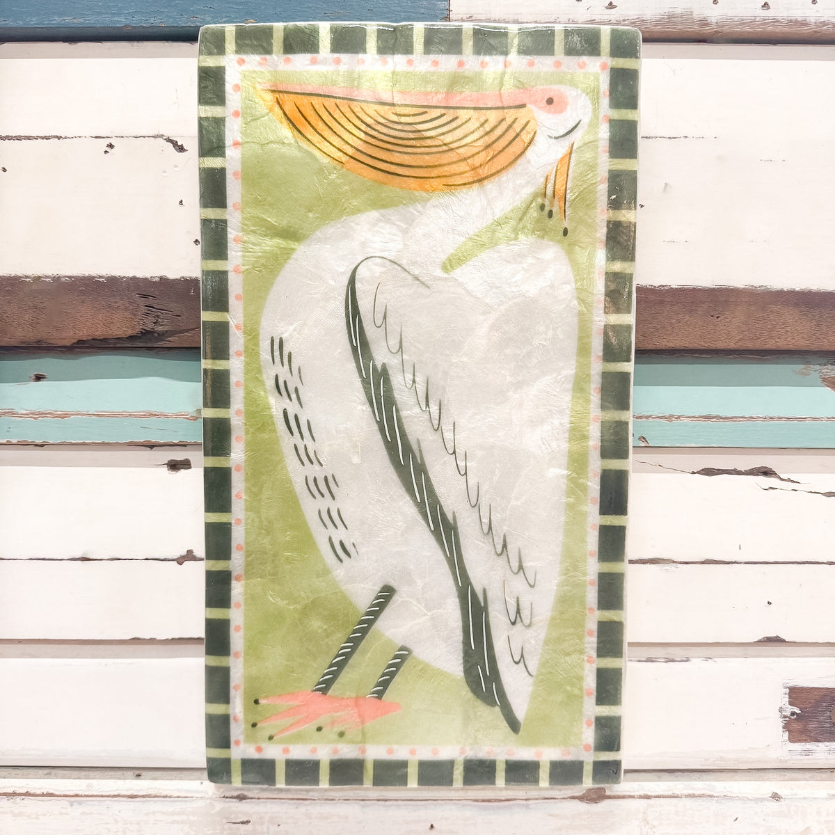 A decorative wall plaque featuring a stylized white pelican with a yellow-orange beak and legs, set against a light green background with a dark green patterned border. The plaque is displayed against a rustic white wooden plank wall.