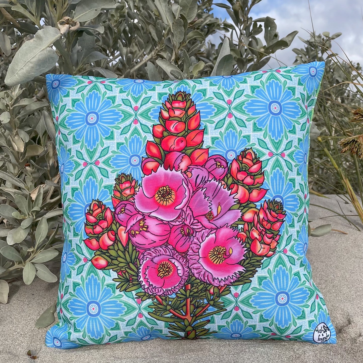 LOST AND FOUND ART CO Featherflower 40cm Cushion. Decorative pillow with pink floral design on a blue floral patterned background, placed outdoors.