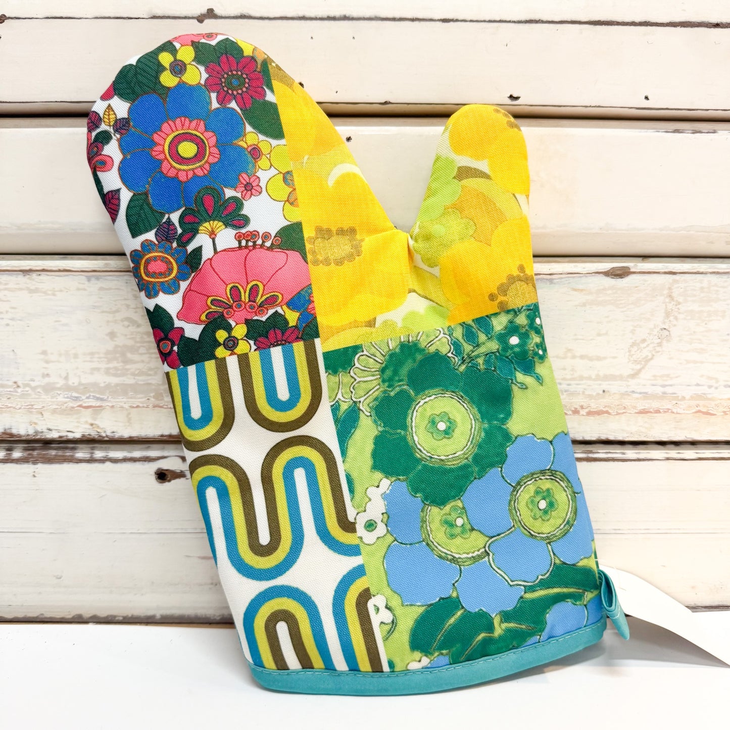 A single fabric oven mitt with a colorful retro patchwork floral and geometric print, displayed against a rustic white wooden plank background.
