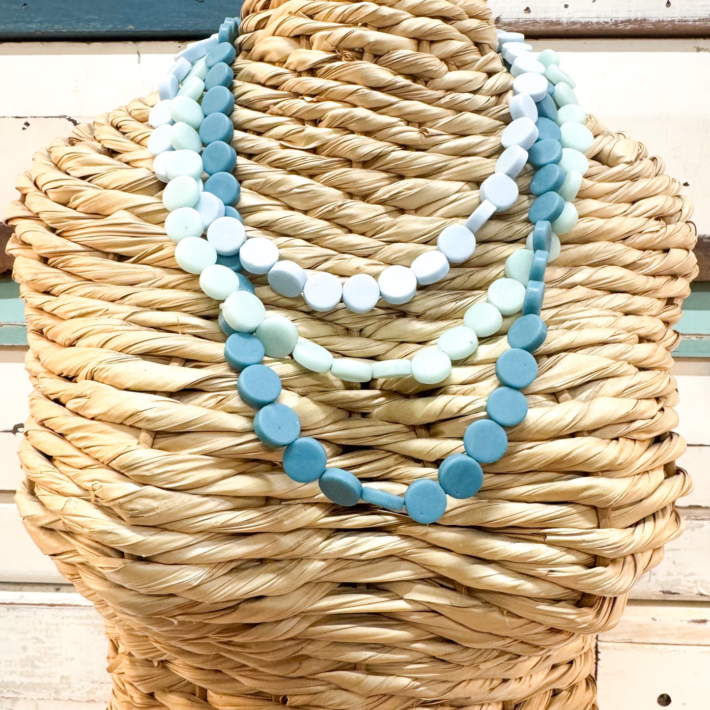 A three-strand beaded necklace set displayed on a woven straw mannequin bust. The strands feature round wooden beads in shades of light blue, medium blue, and white, arranged in three layered styles. The Brightside Store | Pop Necklace - Icey Blue