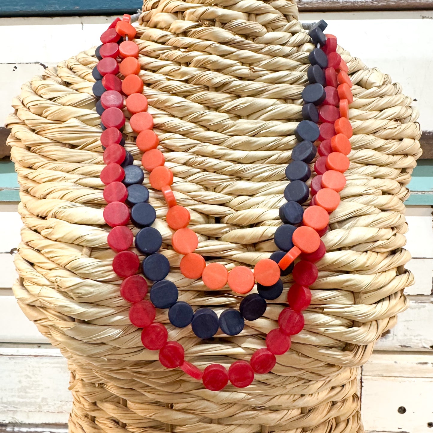Three layered beaded necklaces with round wooden discs in red, orange, and navy blue, displayed on a woven straw mannequin torso.The Brightside Store | Pop Necklace - Sunset Reds