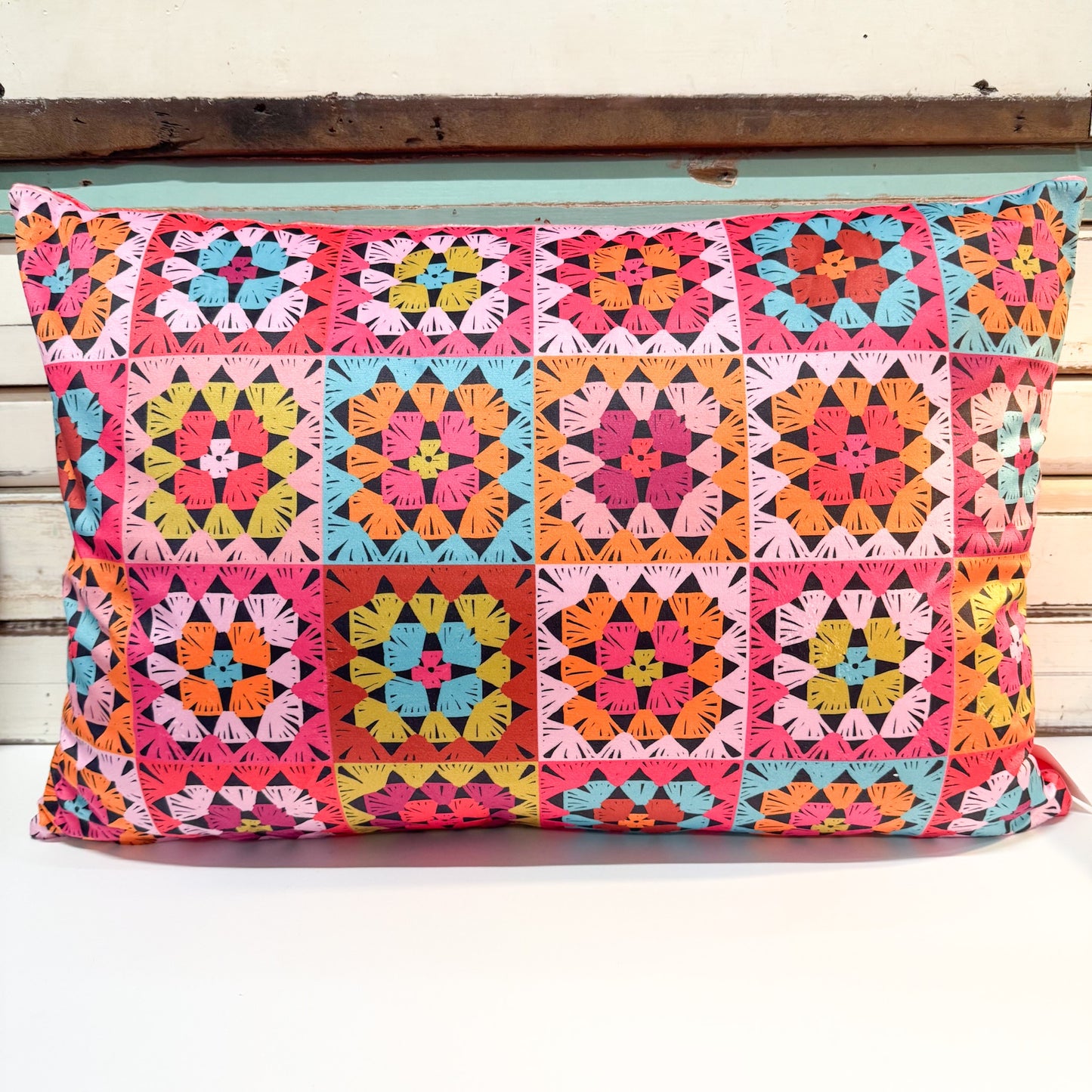 A square decorative throw pillow featuring a vibrant, multicolored geometric patchwork pattern of granny squares in pink, orange, yellow, green, and blue, resting on a white surface against a rustic wooden background.