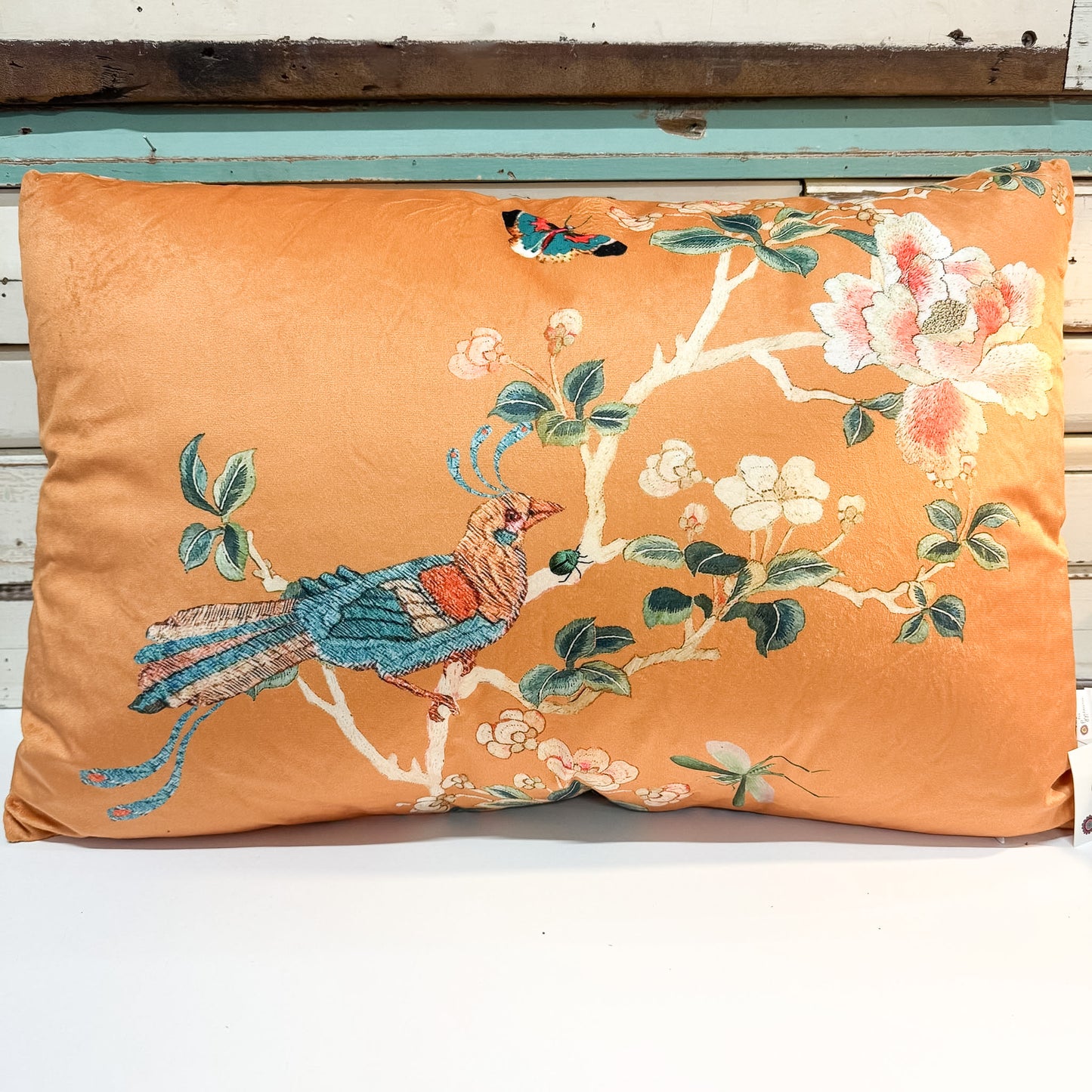 An orange decorative throw pillow featuring a detailed embroidered design of a blue bird, white blossoms, and green leaves on a white wooden background.