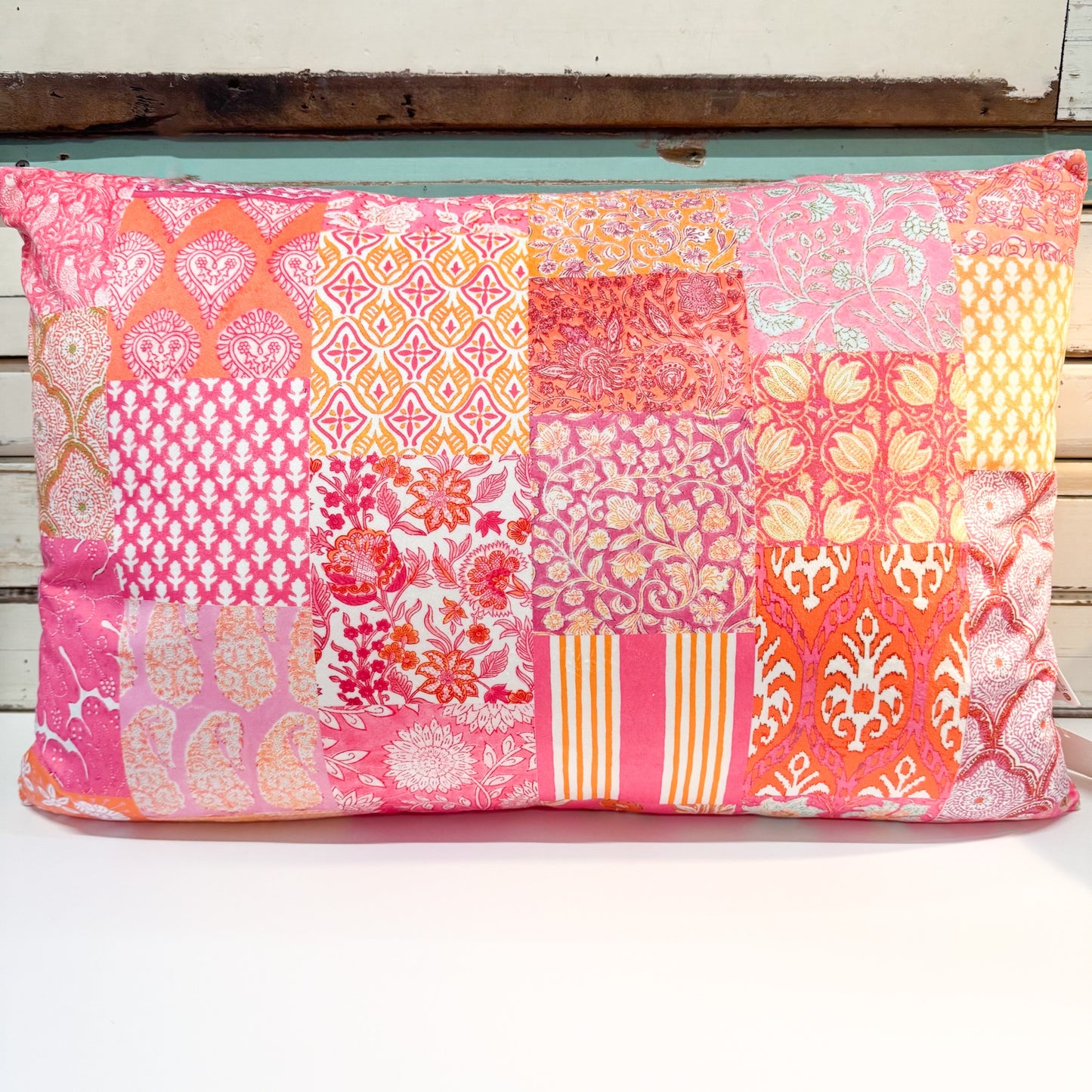 A square decorative throw pillow featuring a vibrant patchwork design of various pink, orange, and yellow fabrics with intricate floral and geometric patterns, resting on a white surface.