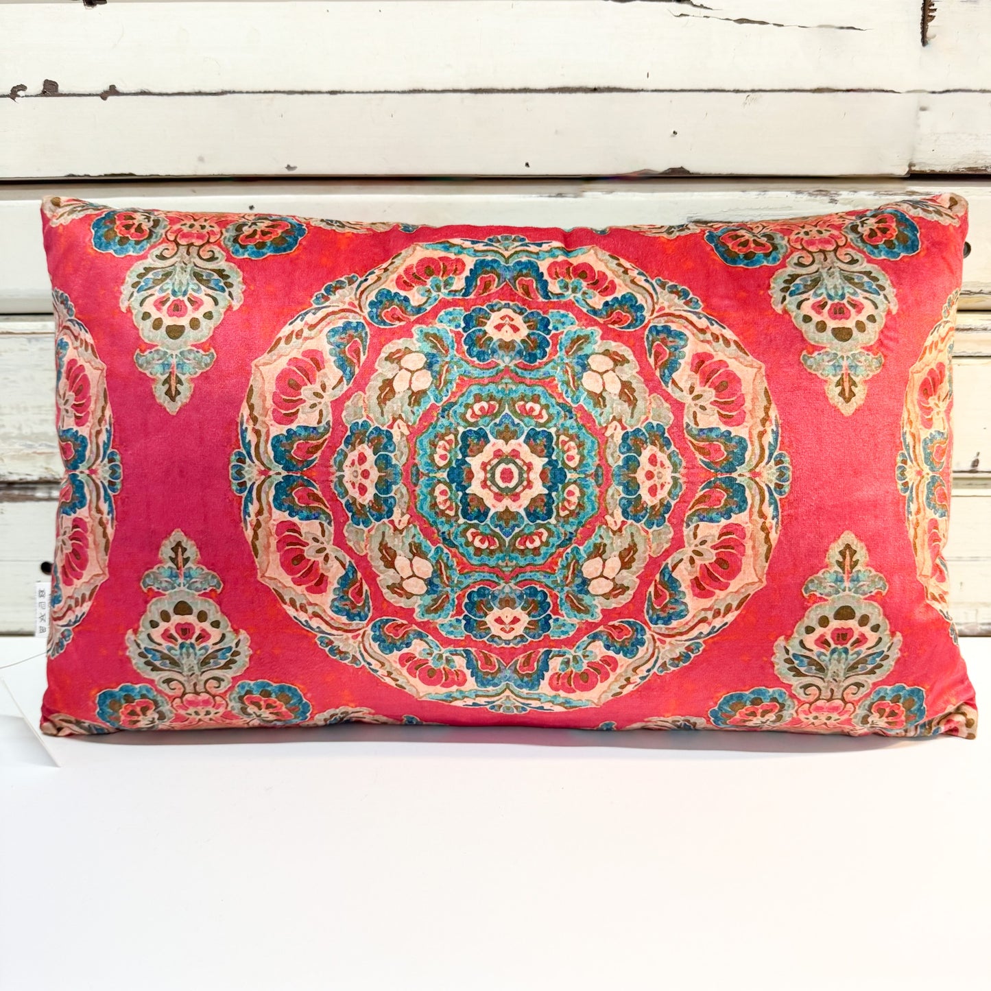 A rectangular throw pillow with a vibrant pink background and an intricate, multi-colored traditional medallion pattern, resting on a white distressed wooden surface.