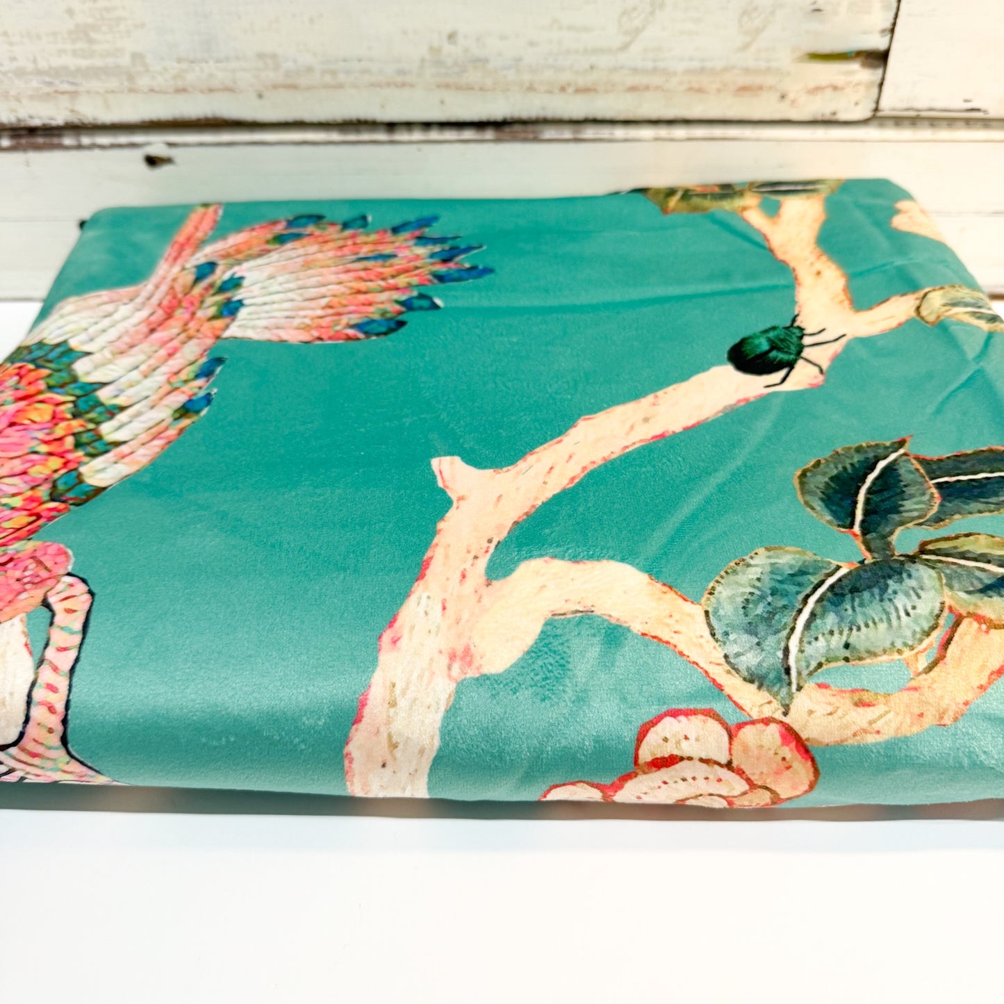 A folded piece of teal silk fabric featuring a traditional Chinese-style print with a phoenix, branch, leaves, and a beetle, displayed on a white wooden surface.