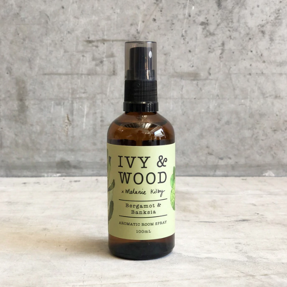A bottle of Ivy & Wood room spray with a spray nozzle, labeled 'Bergamot & Banksia', 100ml volume, on a concrete surface with a textured background.