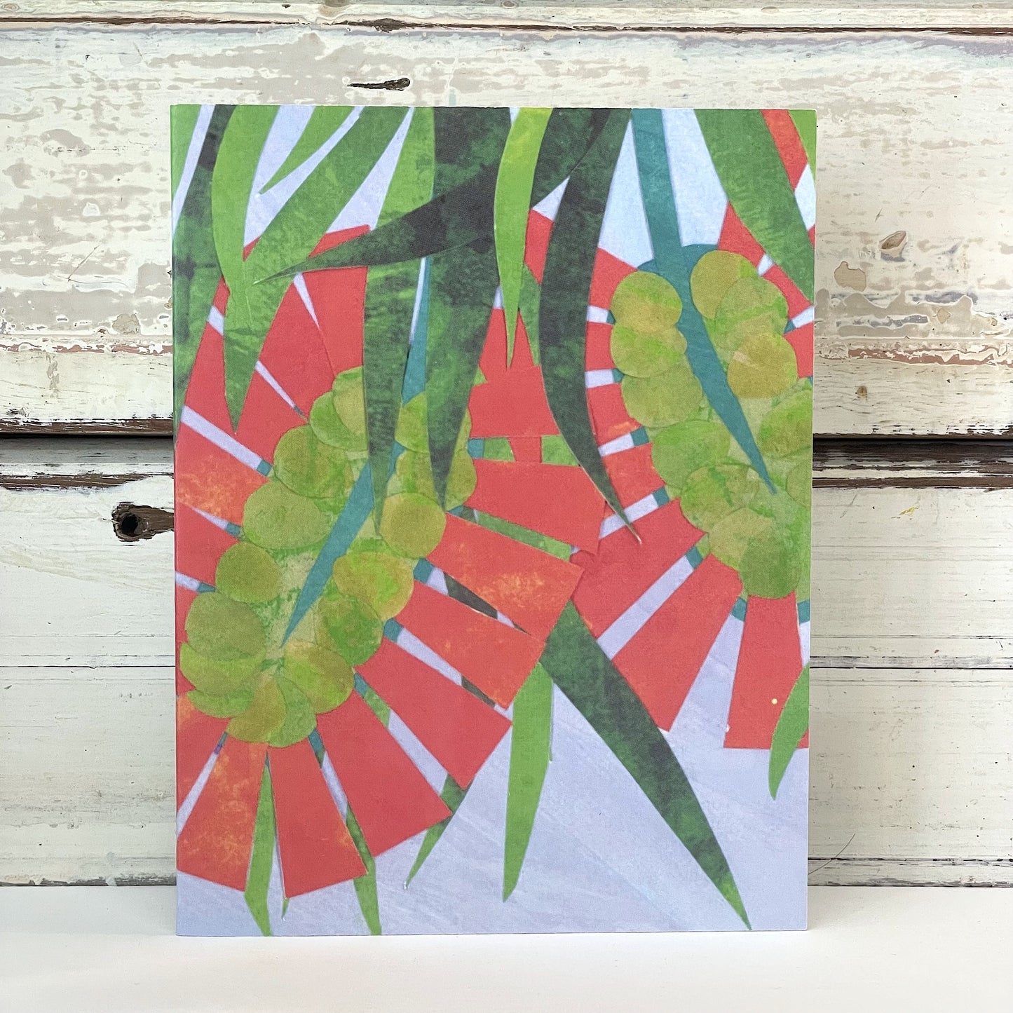 SAMMY JAY BOTTLE BRUSH Card.A colorful greeting card featuring a design with red, green, and blue floral patterns, placed against a wooden background.