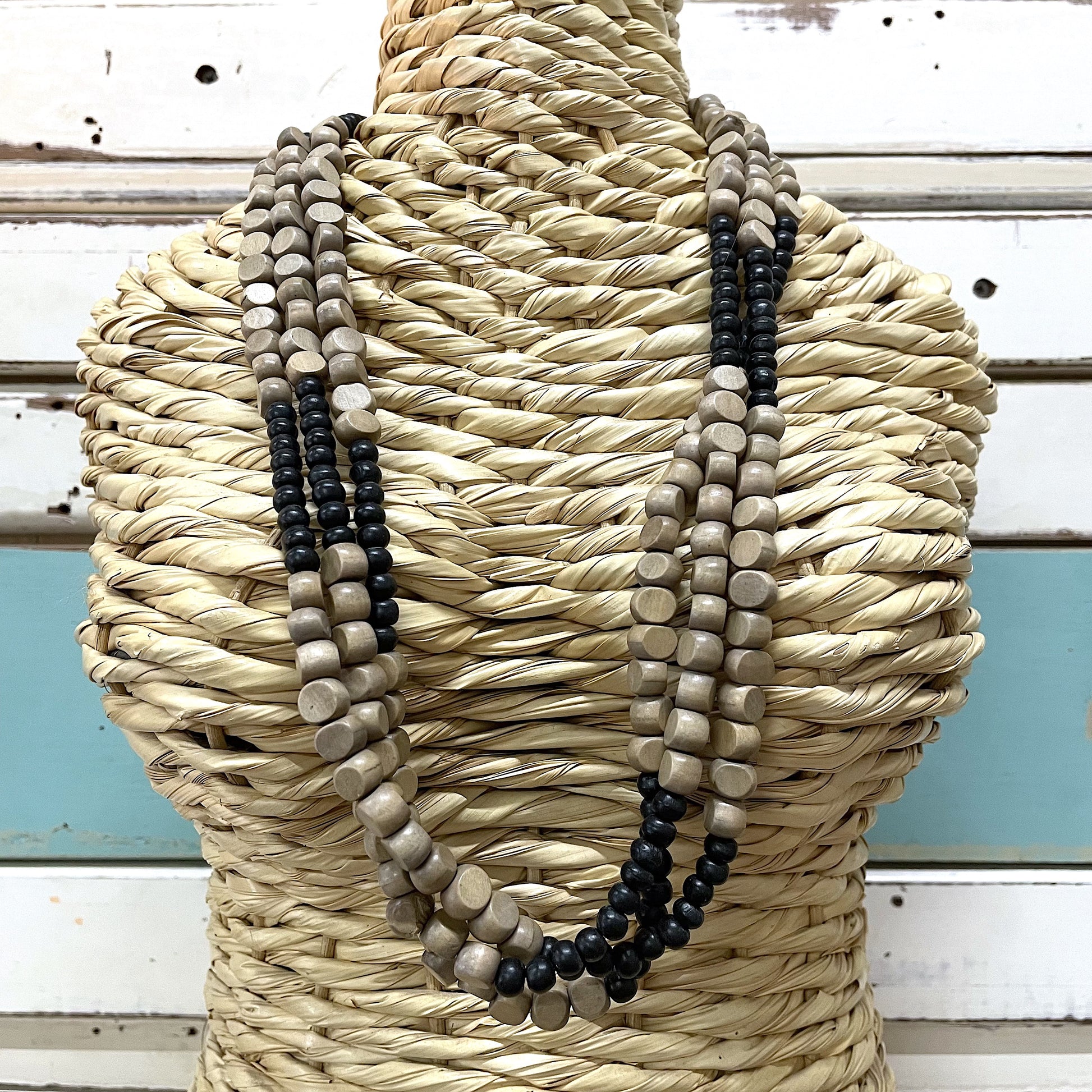 A three-strand wooden bead necklace displayed on a straw mannequin. The beads are in shades of black, beige, and gray.