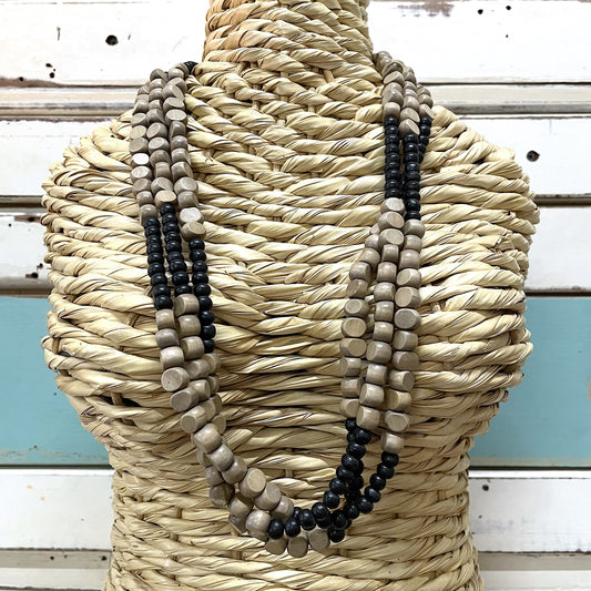 A three-strand wooden bead necklace displayed on a straw mannequin. The beads are in shades of black, beige, and gray.