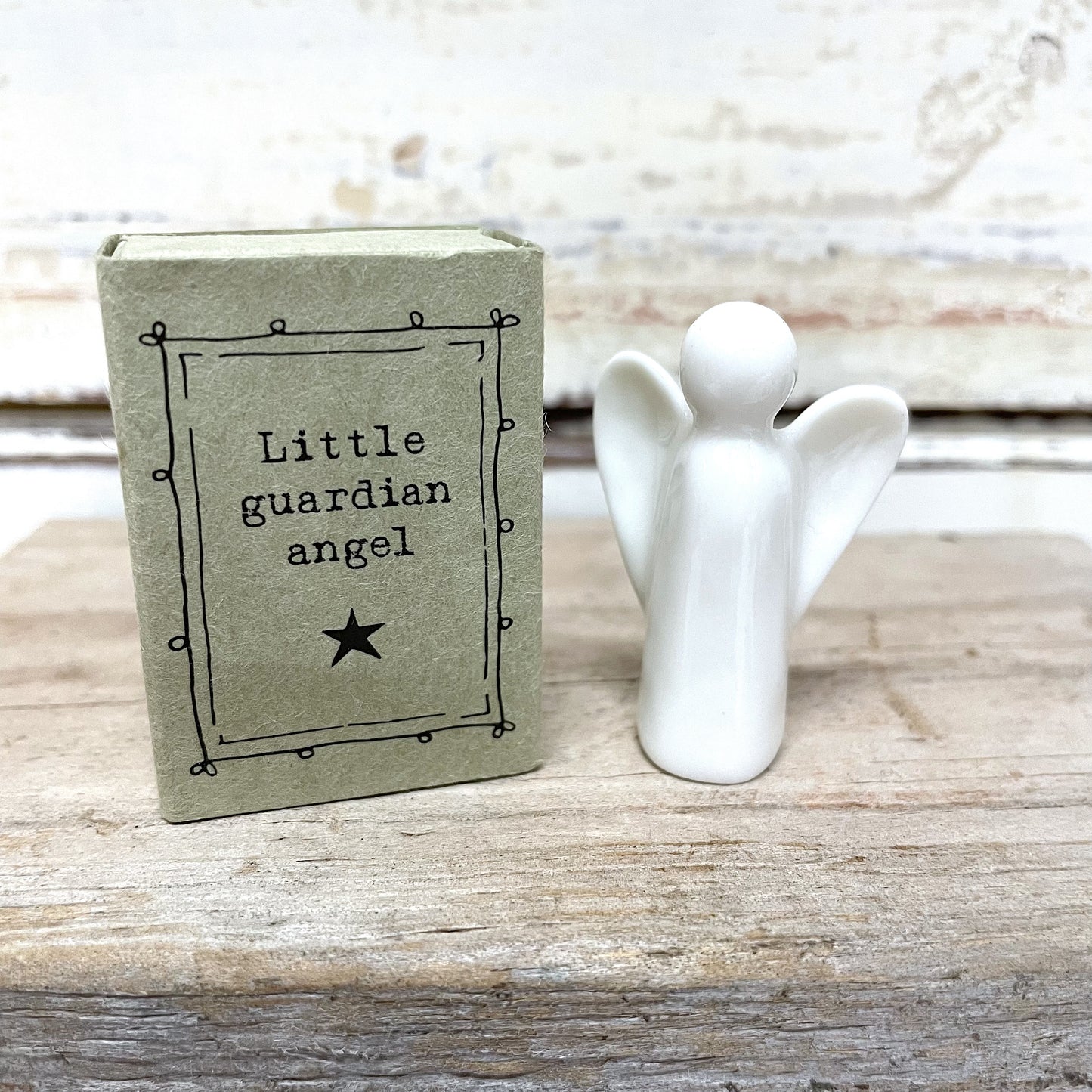 A white porcelain figurine of a little guardian angel sitting on a matchbox box with the text 'Little Guardian Angel' written on it.