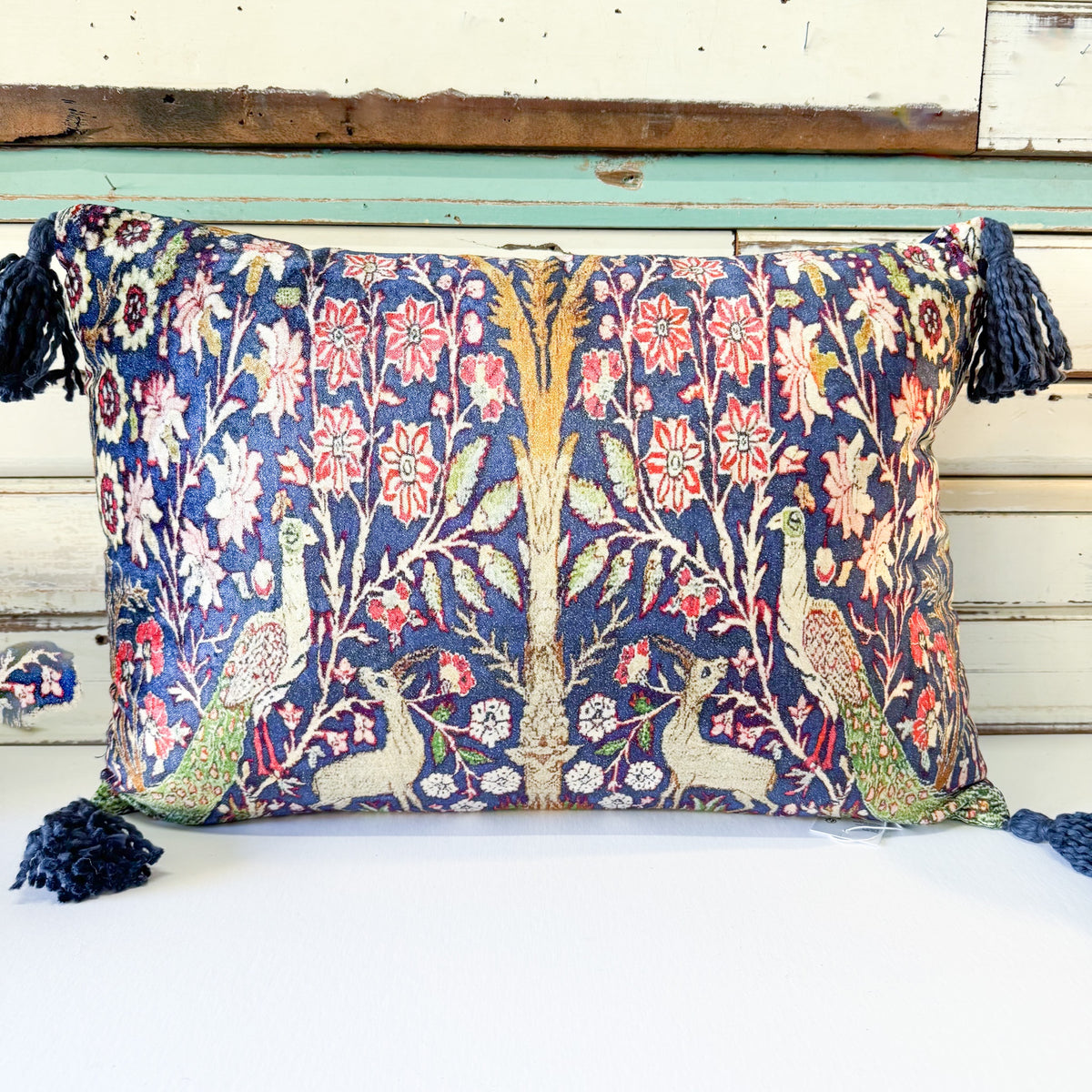 A square decorative throw pillow with a dense floral and peacock tapestry pattern in navy blue, red, green, and cream, finished with dark blue tassels on the sides, set against a rustic white and teal wooden background.