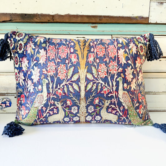 A square decorative throw pillow with a dense floral and peacock tapestry pattern in navy blue, red, green, and cream, finished with dark blue tassels on the sides, set against a rustic white and teal wooden background.