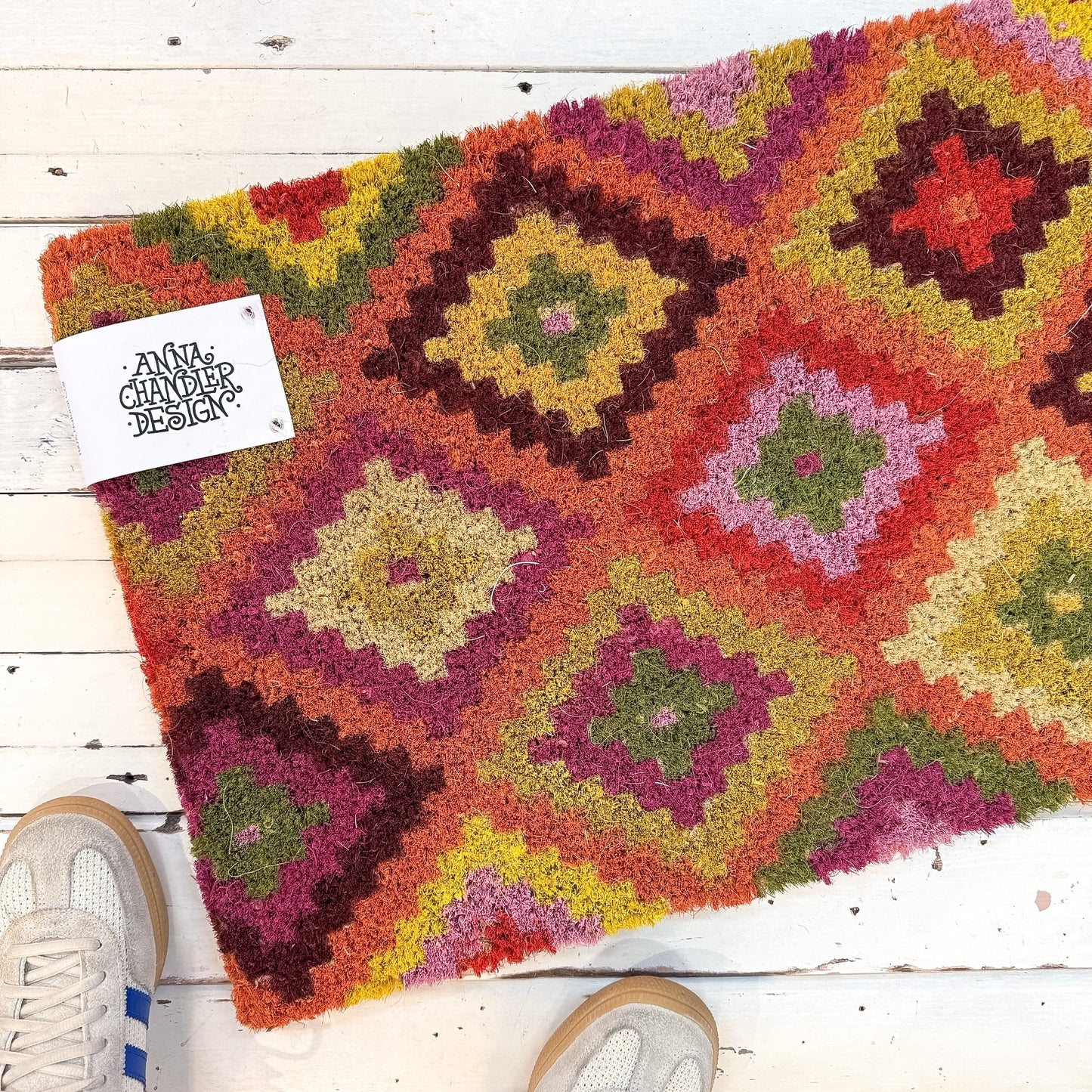 Colourful geometric-patterned rug on a wooden floor with a brand tag. Anna Chandler Doormat - Kelim Orange