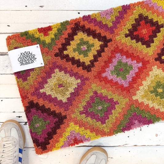 Colourful geometric-patterned rug on a wooden floor with a brand tag. Anna Chandler Doormat - Kelim Orange