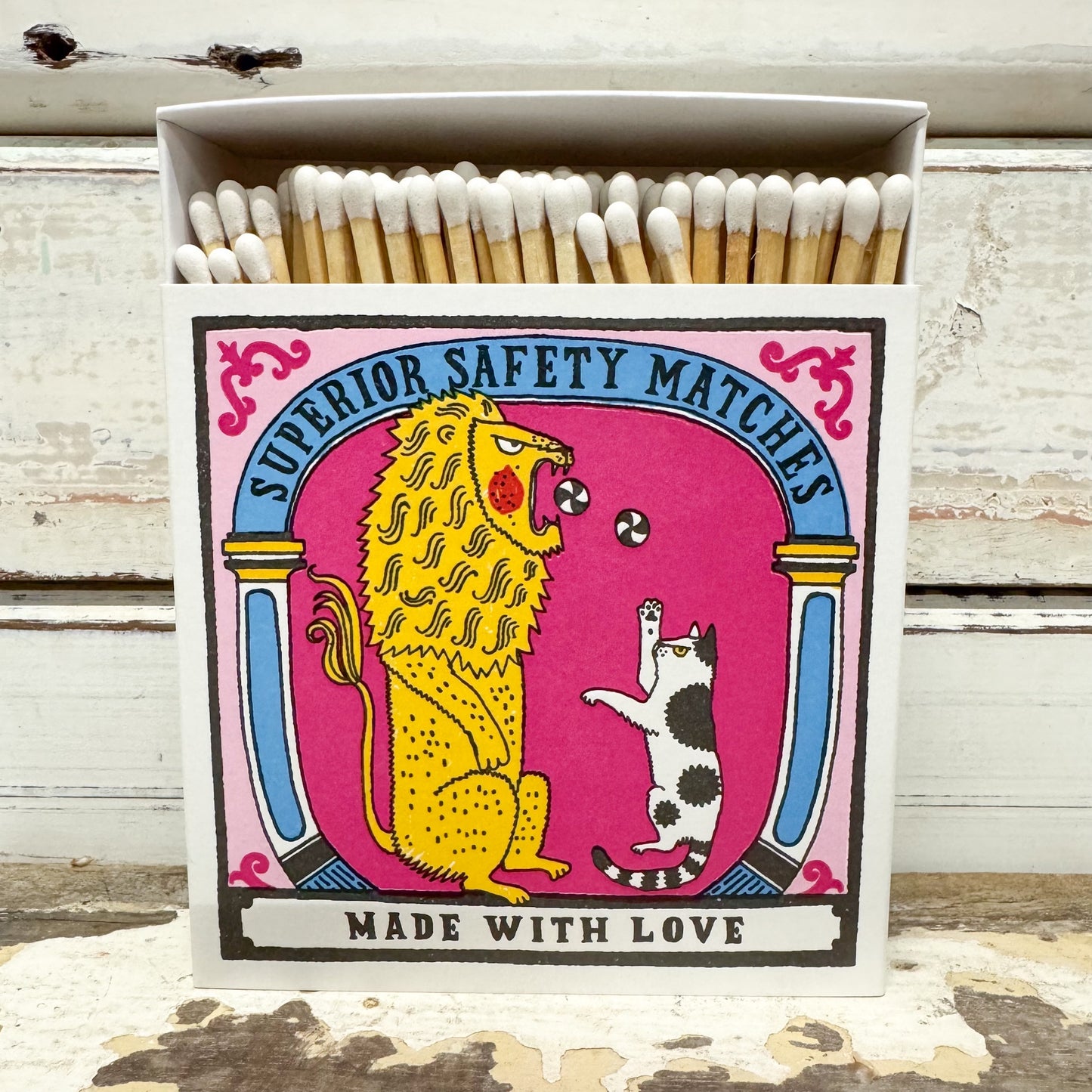 A matchbox of 'Superior Safety Matches' with a colorful illustration of a yellow lion and a black-and-white cat on a pink background, set against a rustic white wooden backdrop.