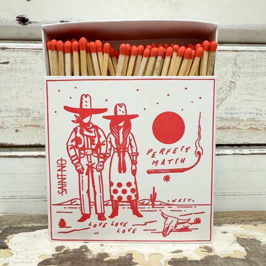 A decorative matchbox filled with wooden matches topped with bright orange-red tips, standing on a weathered white wood surface. The front of the box has a red illustrated design with two cowboy figures, a red circle, and the words 'PERFECT MATCH' and 'SAN ANTO'.