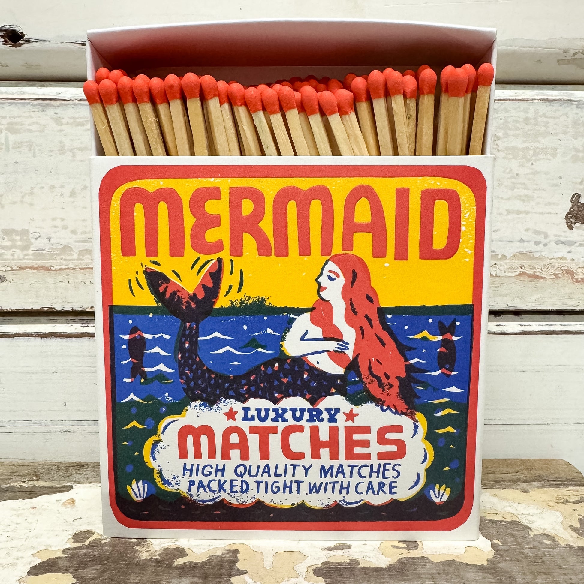 A vintage-style matchbox labeled "MERMAID" filled with wooden matches topped with red tips, set against a weathered white wood background.