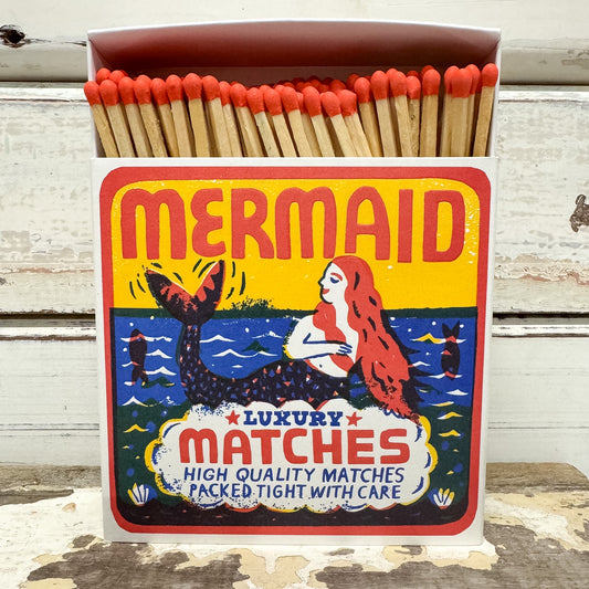 A vintage-style matchbox labeled "MERMAID" filled with wooden matches topped with red tips, set against a weathered white wood background.
