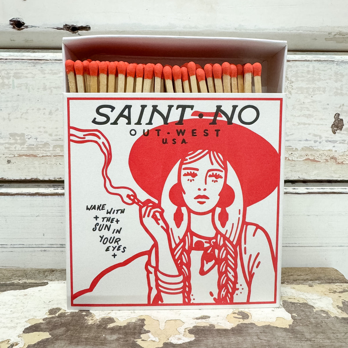 A matchbox-style product with orange match heads and a white box printed with a red illustrated woman and the text 'SAINT-NO OUT-WEST USA' and 'WAKE WITH * THEY JOIN IN YOUR EYES *'. The box is photographed on a rustic white wooden surface.