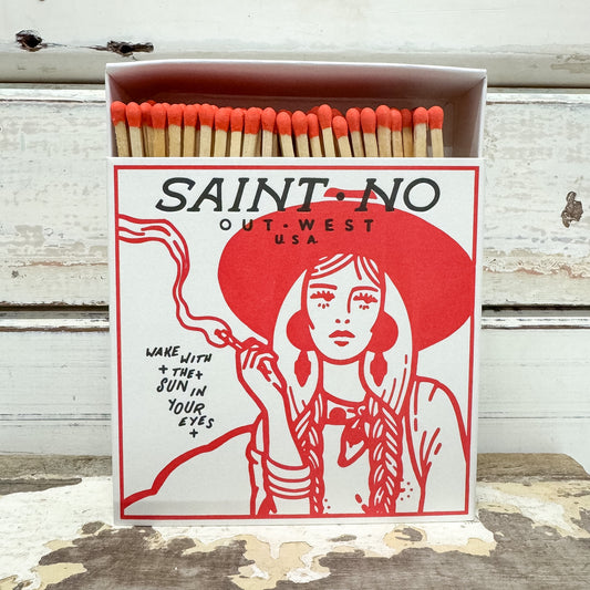 A matchbox-style product with orange match heads and a white box printed with a red illustrated woman and the text 'SAINT-NO OUT-WEST USA' and 'WAKE WITH * THEY JOIN IN YOUR EYES *'. The box is photographed on a rustic white wooden surface.
