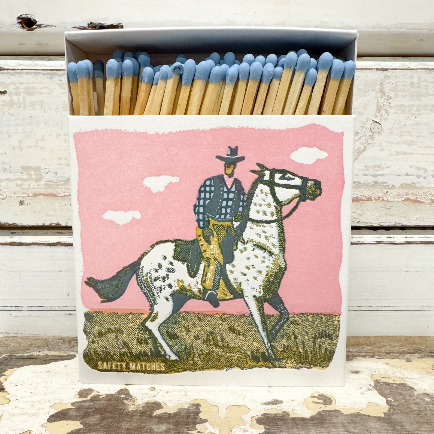 A matchbox filled with wooden safety matches with blue tips, featuring a western-style illustration of a cowboy riding a dappled horse against a pink sky on the front.