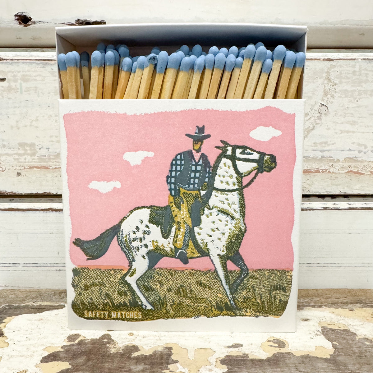 A matchbox filled with wooden safety matches with blue tips, featuring a western-style illustration of a cowboy riding a dappled horse against a pink sky on the front.