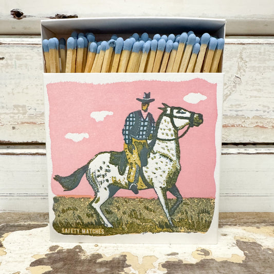 A matchbox filled with wooden safety matches with blue tips, featuring a western-style illustration of a cowboy riding a dappled horse against a pink sky on the front.