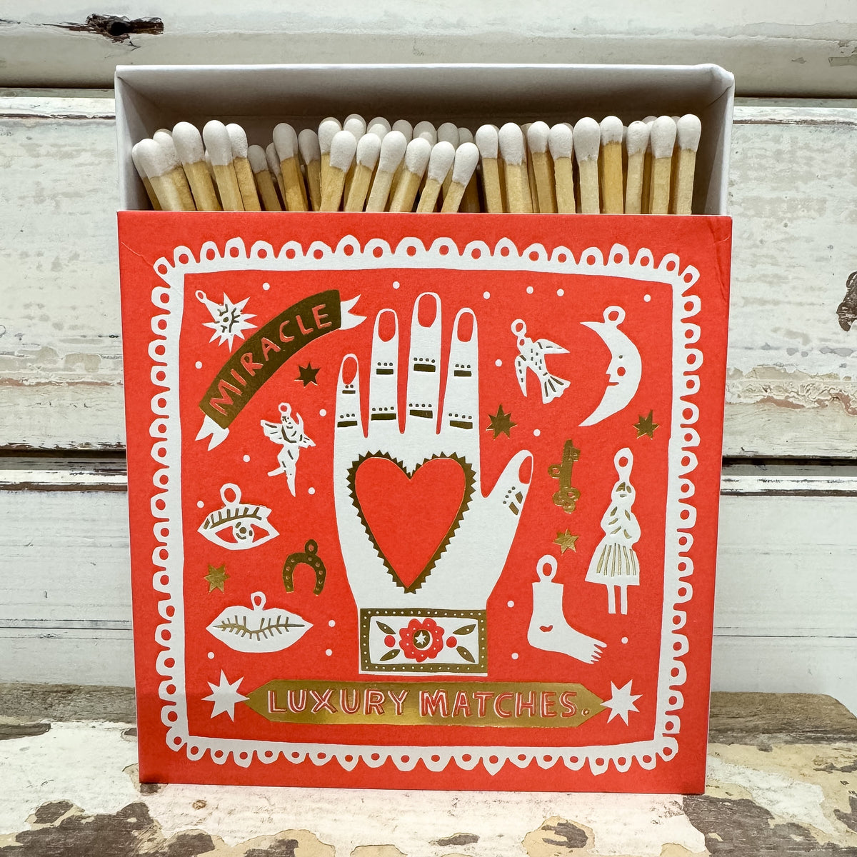 A red decorative matchbox filled with wooden matches, set against a distressed white wooden background. The front of the box features a white illustrated hand with a red heart on the palm, surrounded by whimsical icons like a crescent moon, angel, and stars, with banners reading 'Miracle' and 'Luxury Matches'.