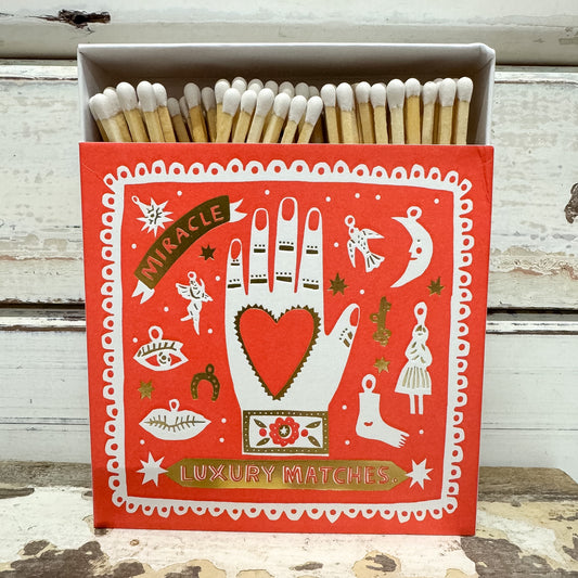 A red decorative matchbox filled with wooden matches, set against a distressed white wooden background. The front of the box features a white illustrated hand with a red heart on the palm, surrounded by whimsical icons like a crescent moon, angel, and stars, with banners reading 'Miracle' and 'Luxury Matches'.