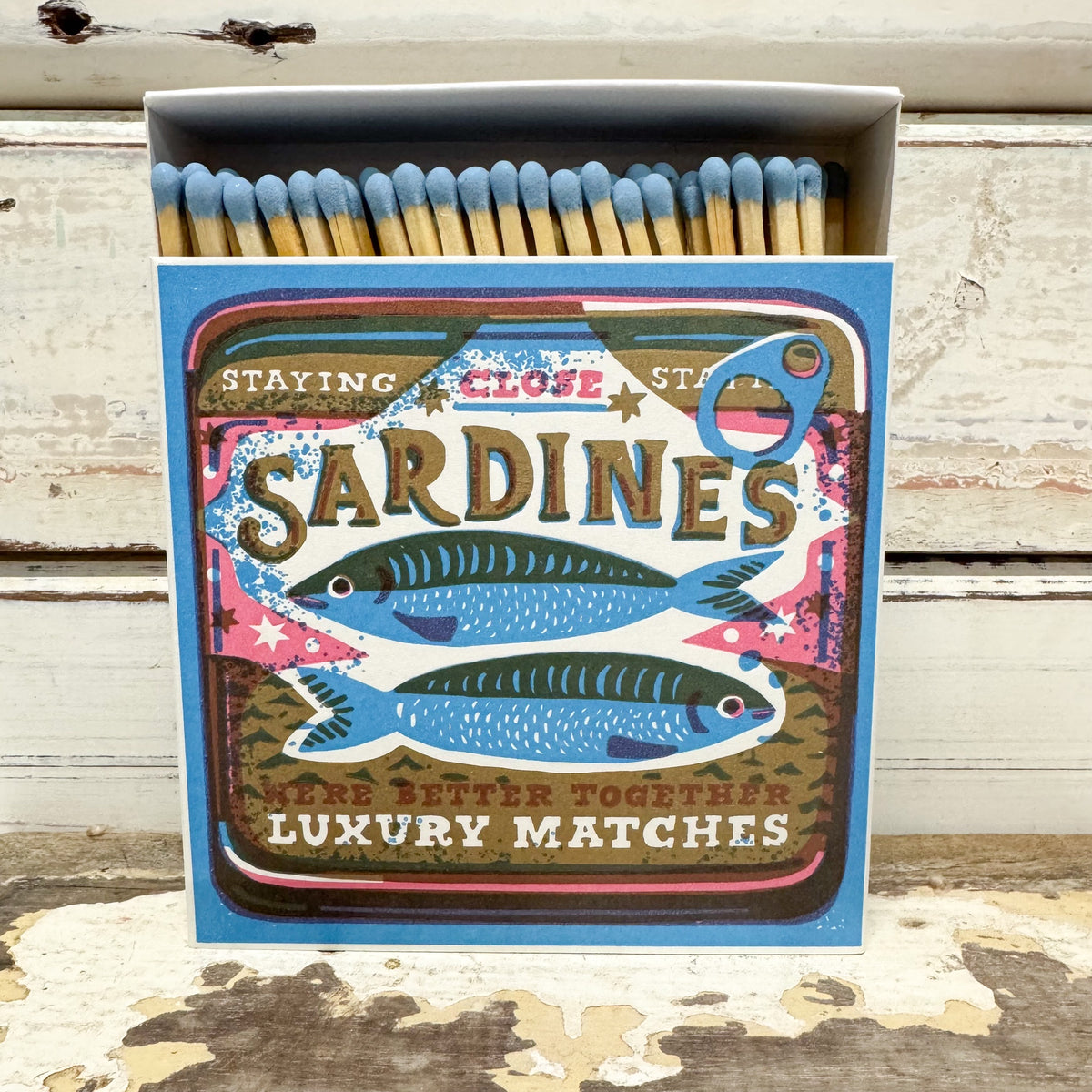 A box of Sardines luxury matches standing open on a distressed white wooden surface. The matchbox features a colorful vintage-style label with two blue sardines, decorative text, and a small bottle opener cutout on the top right.