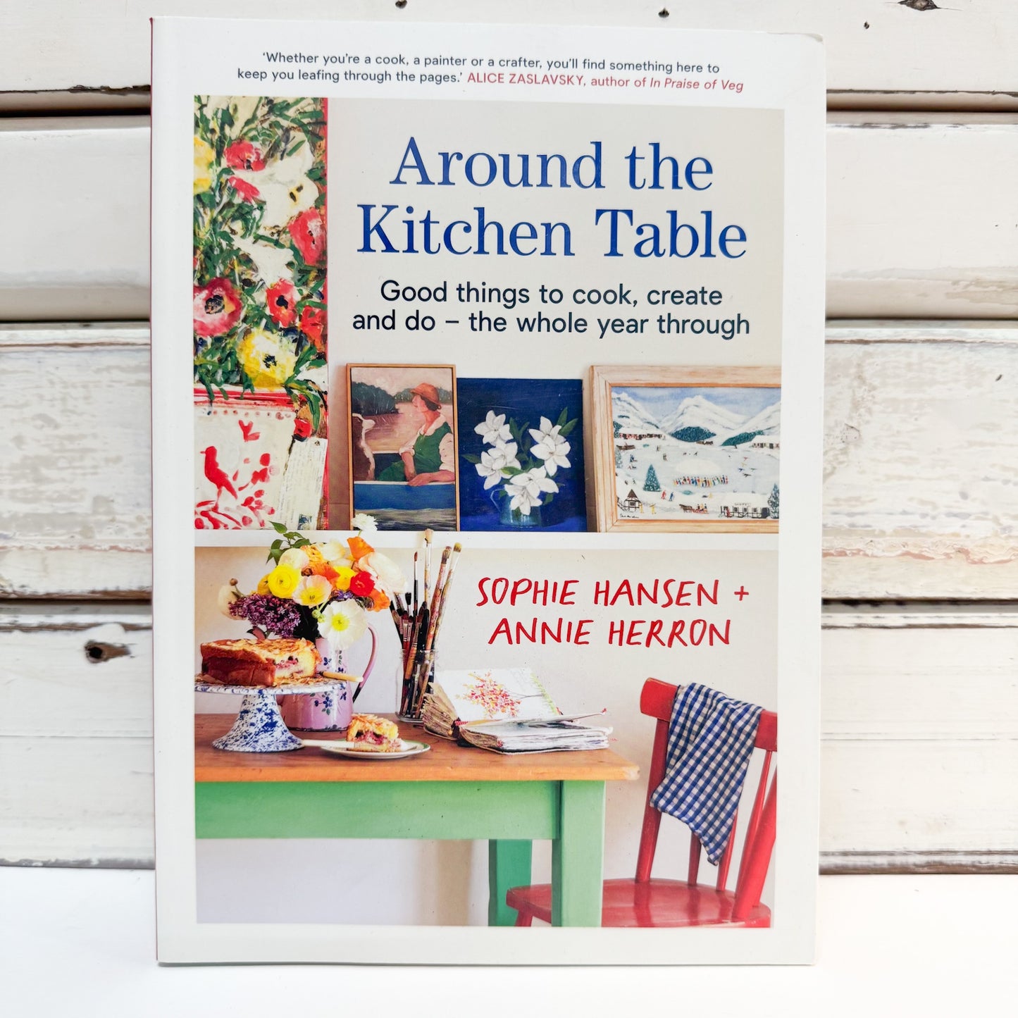 The book 'Around the Kitchen Table' by Sophie Hansen and Annie Herron, displayed upright on a white wooden surface. The cover features a white background with colorful kitchen-themed illustrations, including flowers, paintings, a table, and a red chair.