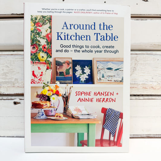 The book 'Around the Kitchen Table' by Sophie Hansen and Annie Herron, displayed upright on a white wooden surface. The cover features a white background with colorful kitchen-themed illustrations, including flowers, paintings, a table, and a red chair.