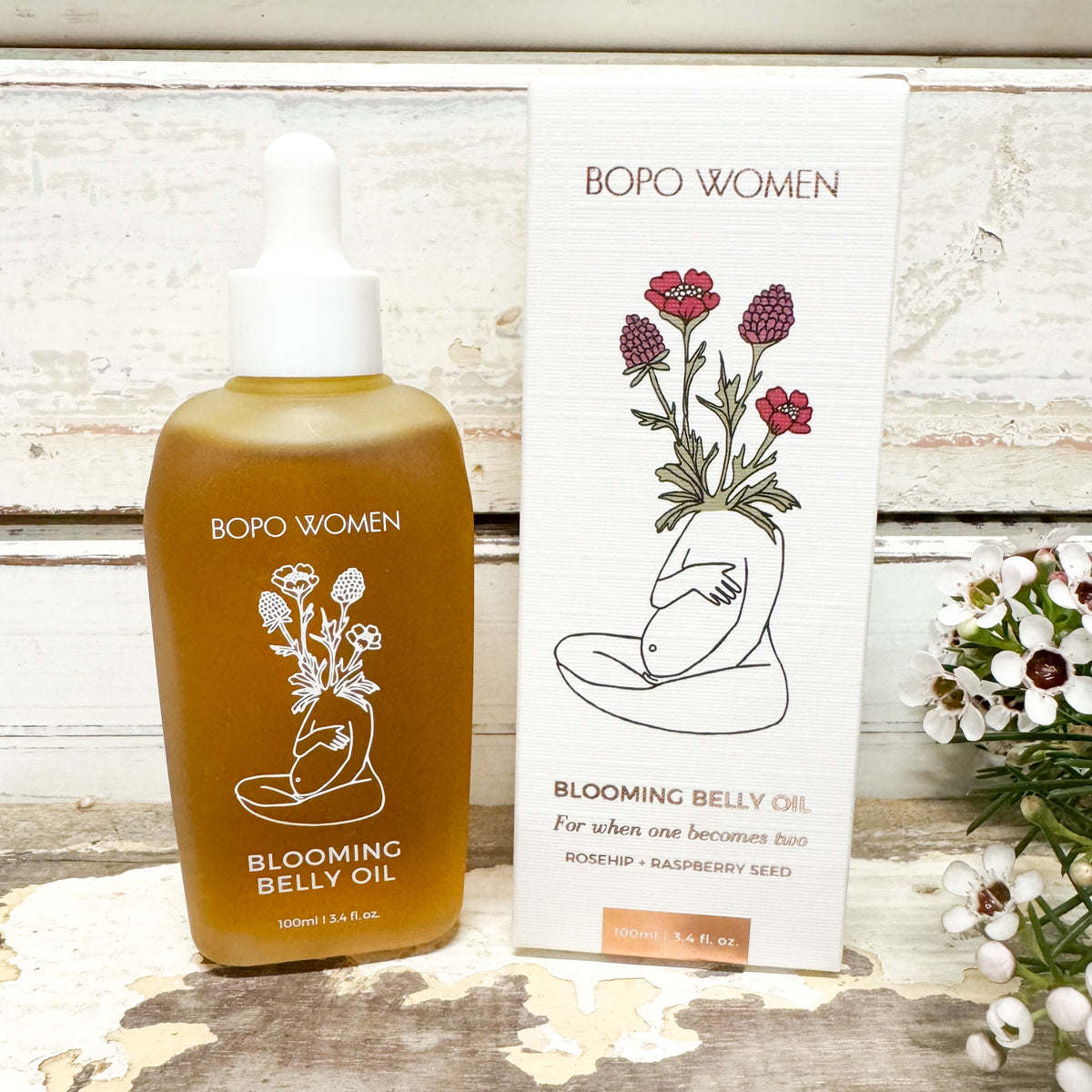 A bottle of Bopo Women Blooming Belly Oil next to its white packaging box, styled on a rustic white wooden surface with small white flowers.