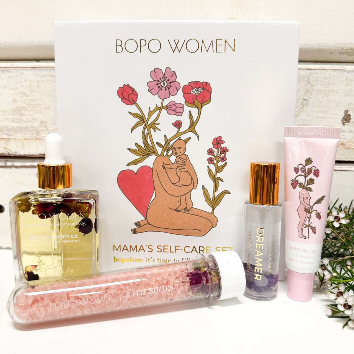 Bopo Women Mama's Self-Care Set featuring a white gift box with a floral illustration, a bottle of rosehip body oil, a pink bath salt tube, a clear bottle with a gold cap containing purple crystals, and a pink lotion tube, arranged on a white surface in front of the box.