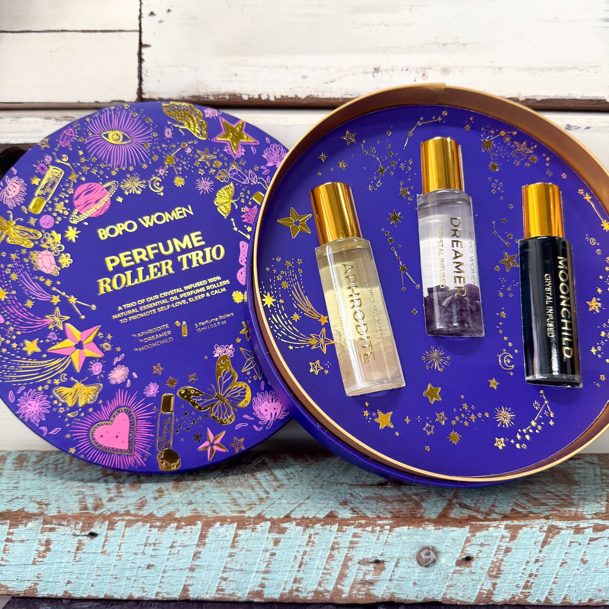 Boro Women Perfume Roller Trio gift set with three roller bottles displayed inside an open round tin next to its illustrated lid, set on a rustic wooden surface.