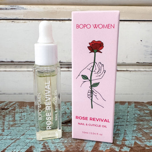 A bottle of Bopo Women Rose Revival Nail & Cuticle Oil next to its pink packaging box, displayed on a rustic wooden surface with a whitewashed wood background.
