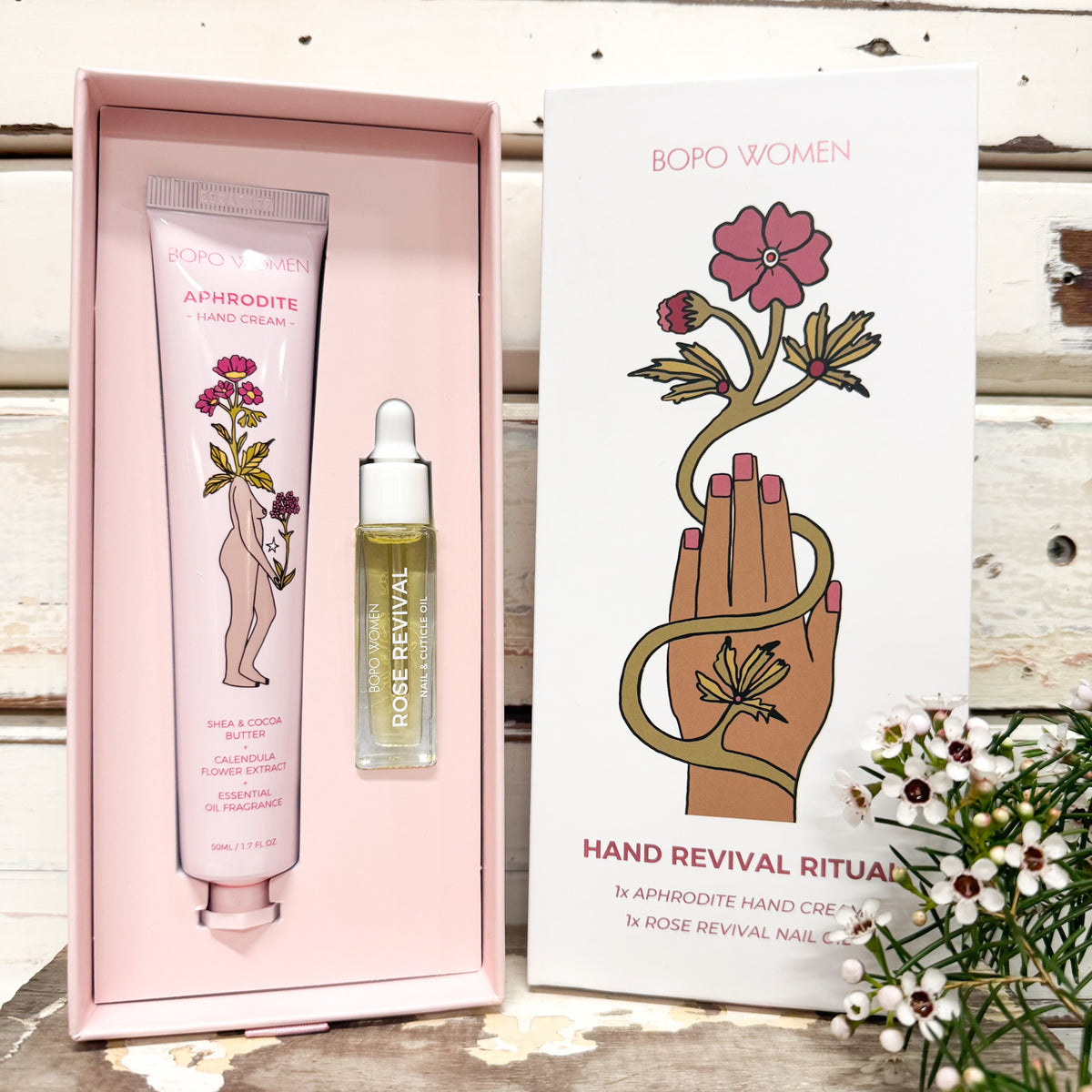 BOPO WOMEN Hand Revival Ritual gift set featuring a pink hand cream tube, a rose revival oil dropper bottle, and a white illustrated box on a rustic wood background.