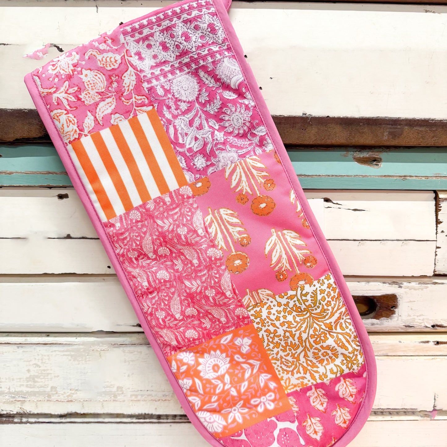 A pink patchwork quilted oven mitt with orange and white patterns, laid diagonally on a distressed white wooden surface.