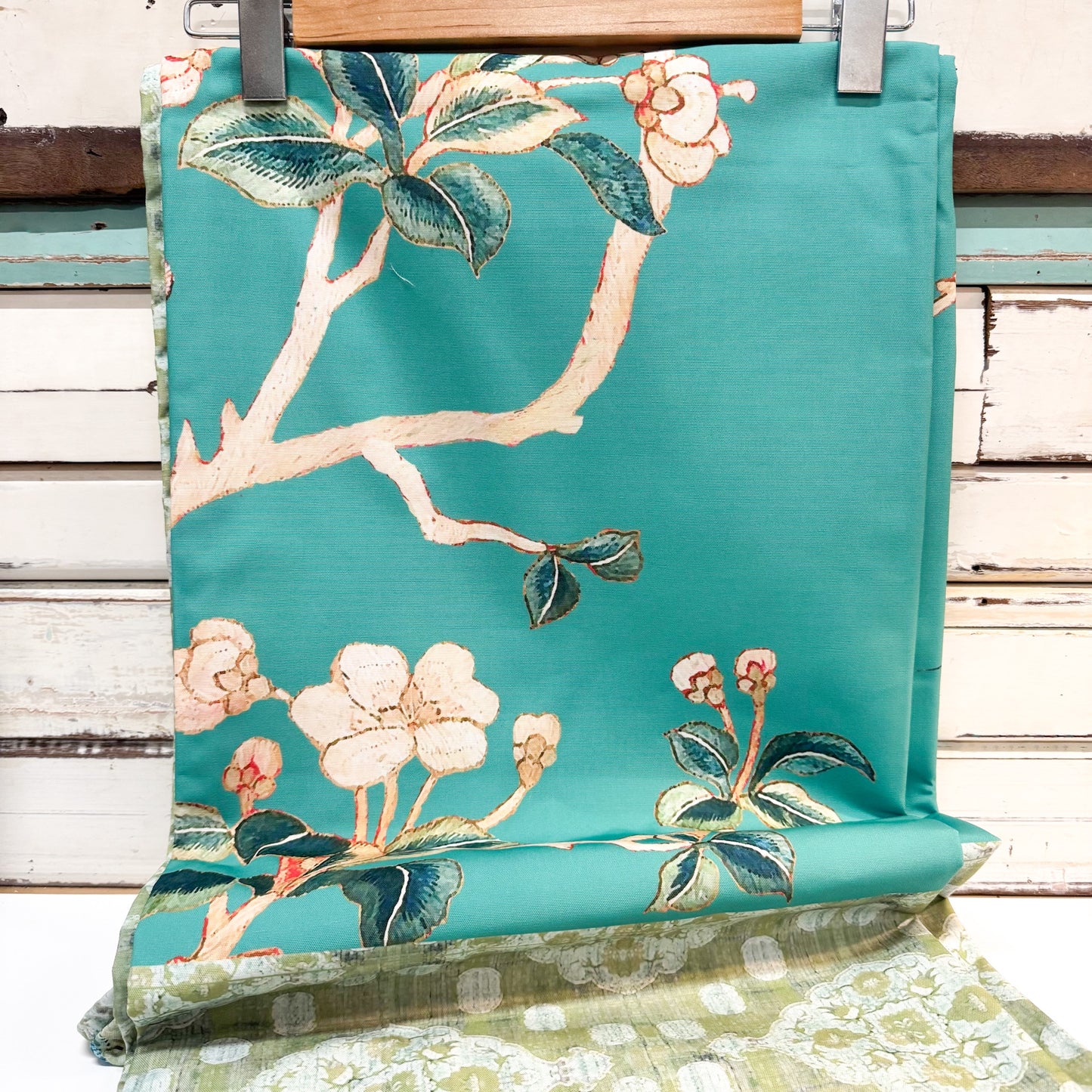 A turquoise outdoor chair cushion with an embroidered floral branch pattern, hanging on a wooden hanger against a rustic white-painted wood plank background.