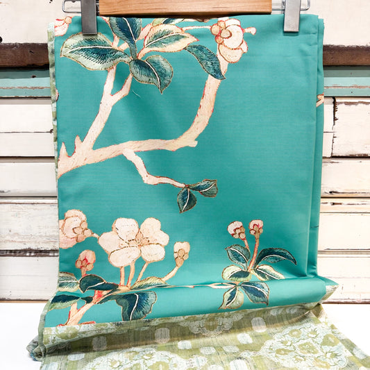 A turquoise outdoor chair cushion with an embroidered floral branch pattern, hanging on a wooden hanger against a rustic white-painted wood plank background.