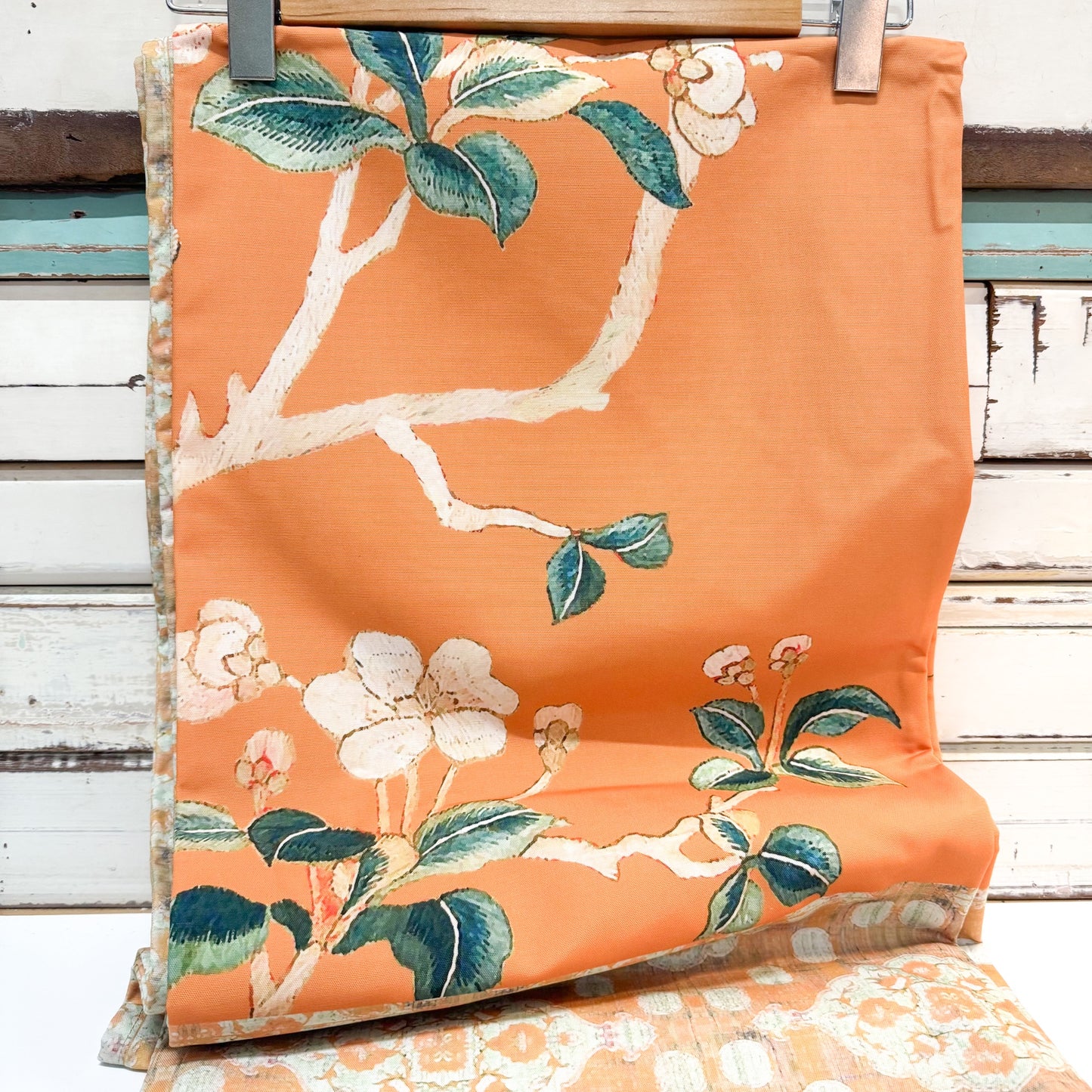 An orange fabric cushion cover featuring a white cherry blossom branch and leaf pattern, hanging on a wooden hanger against a distressed white wood panel background.